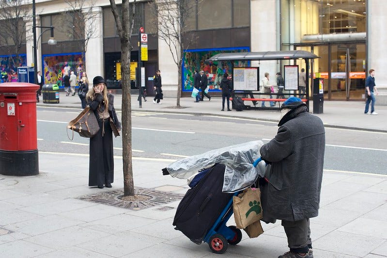 A woman in nice clothes and carrying a designer handbag talks on a mobile phone while a homeless man pushes a plastic covered dolly along the same sidewalk. A woman in nice clothes and carrying a designer handbag talks on a mobile phone while a homeless man pushes a plastic covered dolly along the same sidewalk.