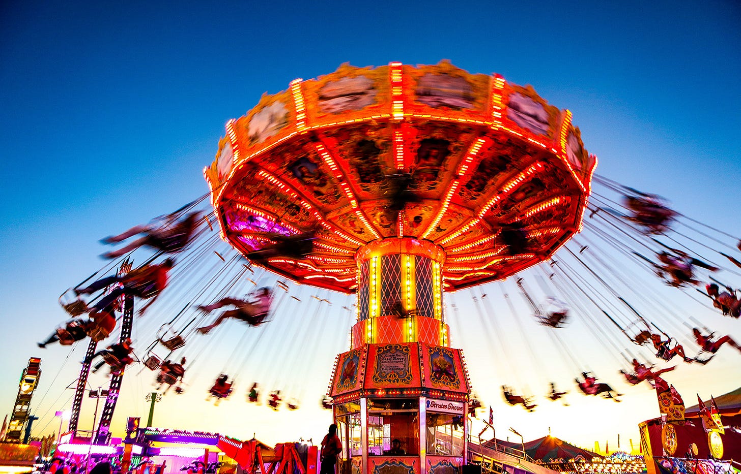 Spinning amusement park ride with figures sitting on chairs suspended by long chains. The ride spins quickly and the figures appear blurred by its speed.