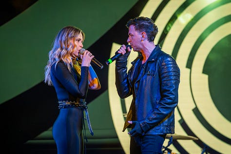 Alina Crișan & Keo doi artiști cântă împreună pe scenă, față în față, fiecare ținând câte un microfon. Artista poartă o ținută neagră, iar artistul poartă o geacă din piele. Fundalul scenei are elemente grafice luminoase în tonuri de verde și galben, iar atmosfera este specifică unui concert live.