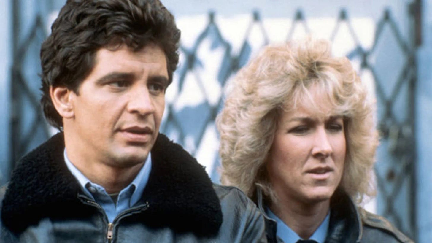 Hill Street Blues still; a male and female cop standing againt a fence, doing nothing and in no particular danger, which is unusually true to life for a cop show