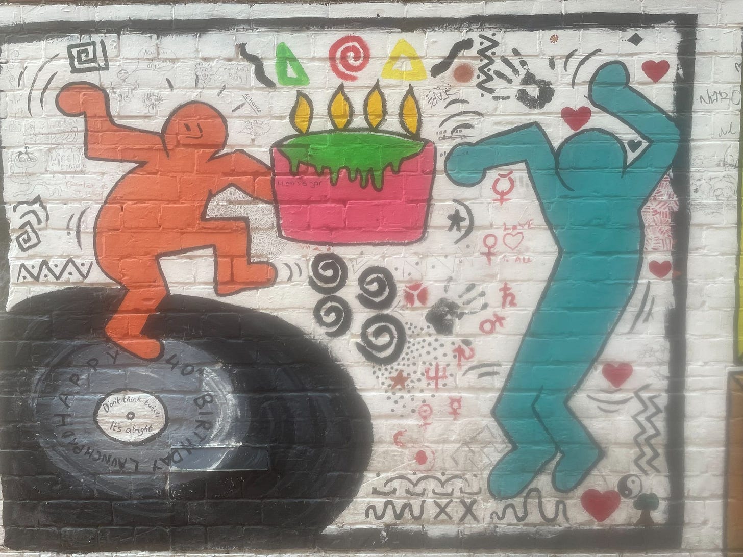 Graffiti art in the style of Keith Haring showing two figures (one orange and one turquoise) dancing on a black vinyl record whilst holding a pink cake with green icing and four lit candles