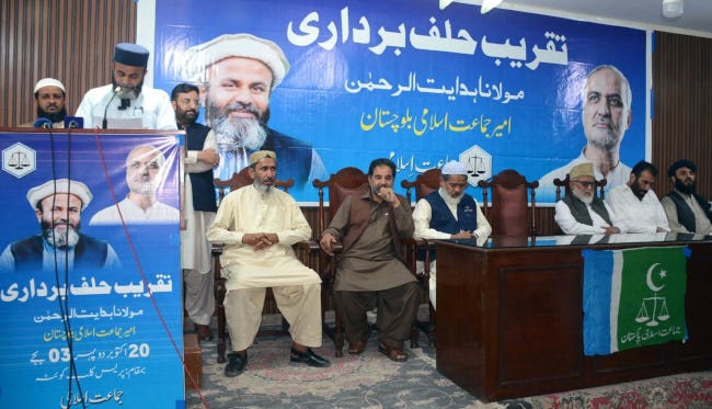 Le Maulana debout à un pupitre, face à un micro et devant une affiche de campagne, prête serment sur une estrade de bois