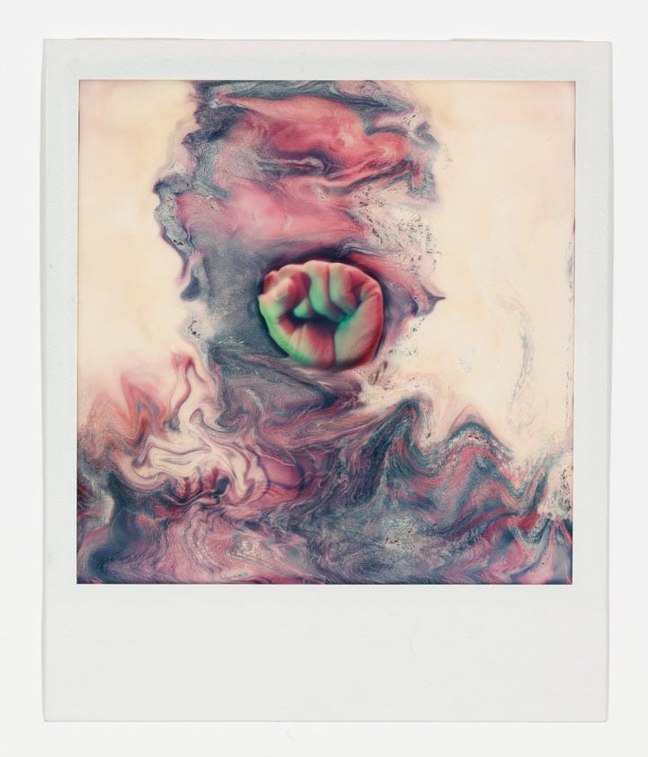 A Polaroid photo featuring an abstract swirl of colors, predominantly in shades of pink, red, and green, with a creamy background. The central focus is a distorted circular shape with a mix of these colors, resembling a marbled pattern.