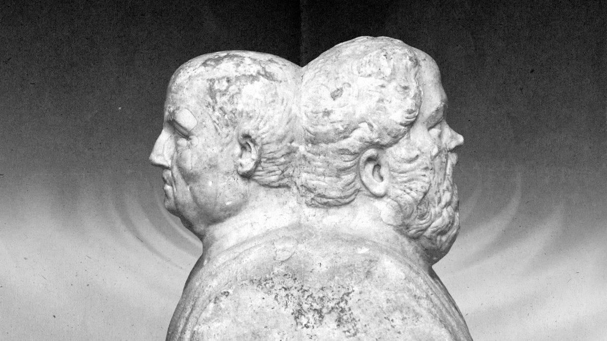 A stone sculpture of two male heads positioned back-to-back, each facing opposite directions, against a plain background.