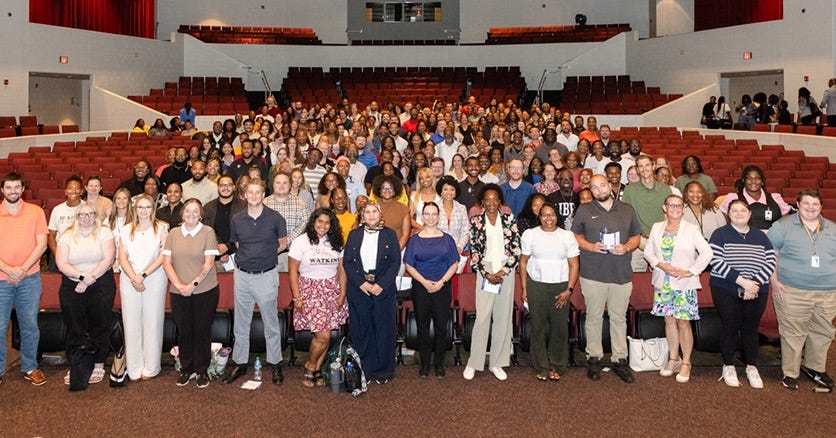 Group photo of hundreds of employees gathered inside a large auditorium during a new employee orientation event, standing together in front of rows of red theater seats. Group photo of hundreds of employees gathered inside a large auditorium during a new employee orientation event, standing together in front of rows of red theater seats.