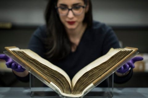 A woman wearing protective gloves prepares to lift the Domesday Book from a display