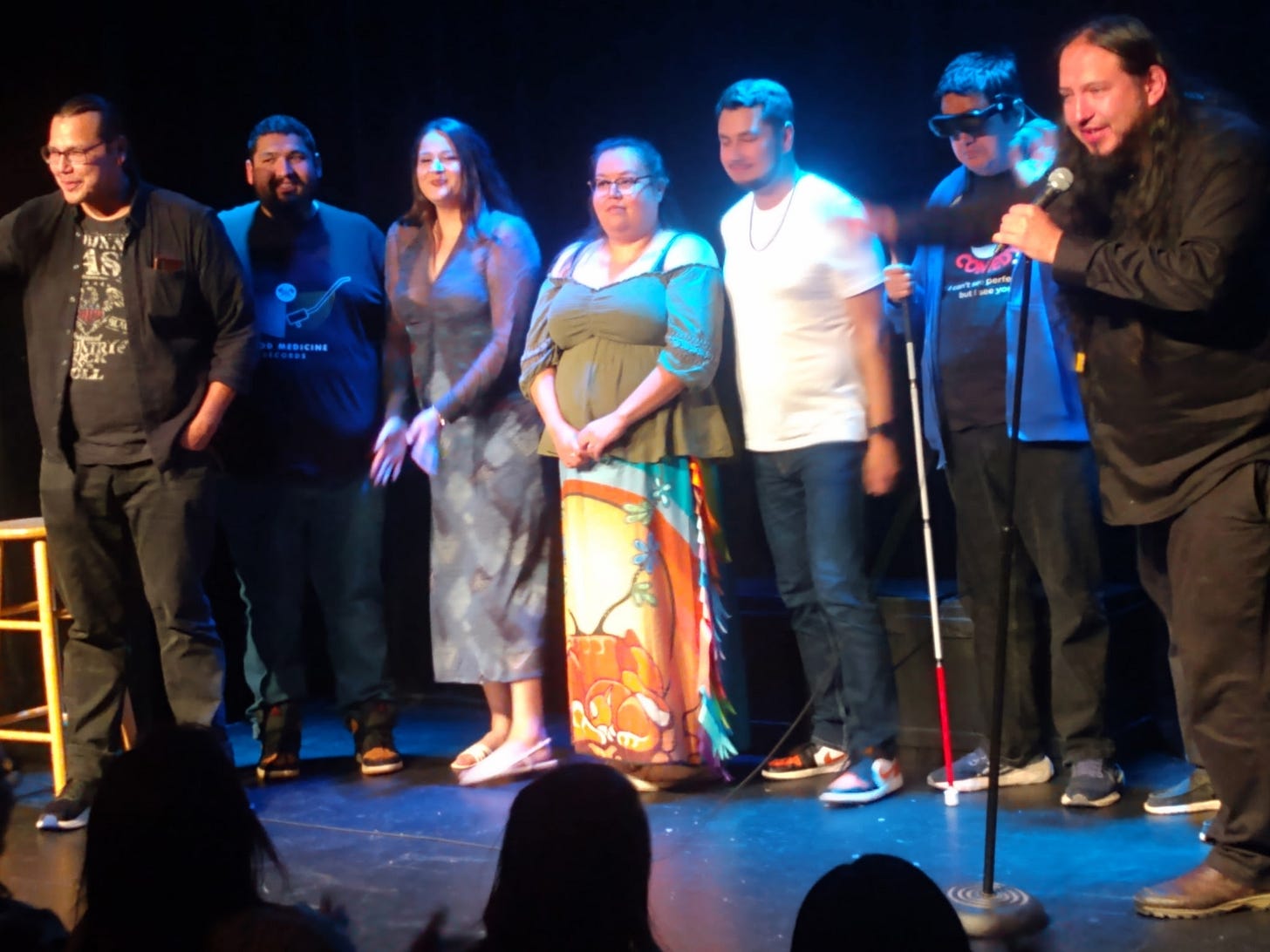 Seven performers stand together on stage at the Grindstone Theatre during the It Must Be Nice Indigenous Comedy Tour. The comedians are lined up shoulder to shoulder under blue stage lighting while the host on the far right speaks into a microphone and gestures toward the group. J.R. stands second from the right wearing dark eSight smart glasses, a black comedy T-shirt, and holding his white cane. Audience silhouettes are visible at the bottom of the image, creating the feel of a live comedy show.