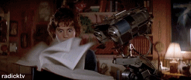 Fermo immagine dal film "Corto Circuito" in cui il robot Numero 5 sfoglia freneticamente un libro sotto lo sguardo sbigottito e sgranato della co-protagonista interpretata da Ally Sheedy.