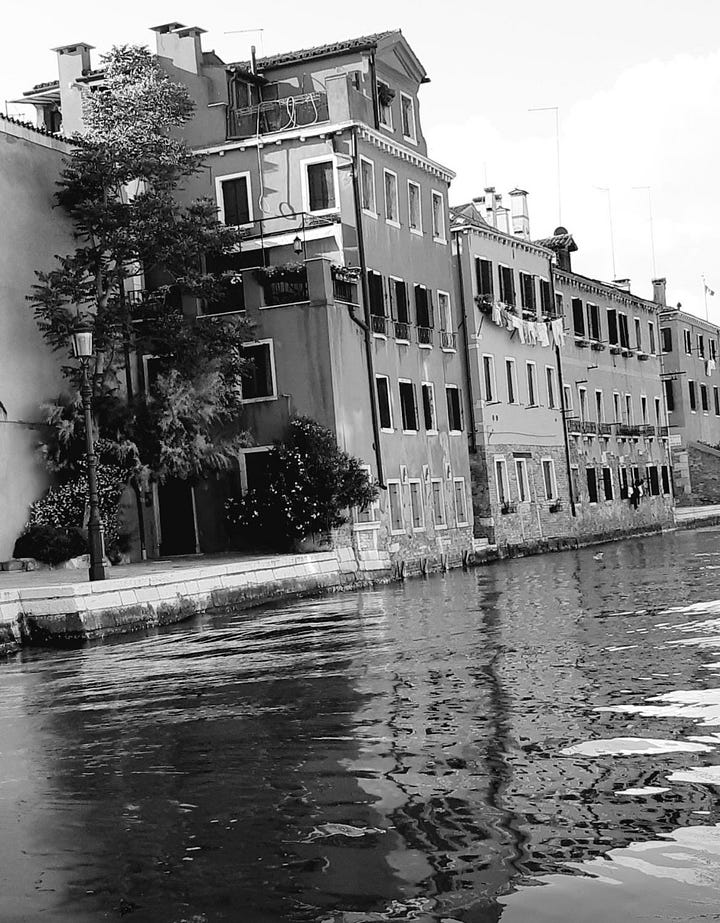 Image 1: A picturesque stone bridge crossing a Venetian canal at sunset, with people walking across and reflections of historic Italian architecture shimmering in the blue water. A small boat is docked on the left, and a terracotta building with white trim stands on the right. Photo taken in Venice, Italy. Image 2: Outdoor summer gathering in an Italian backyard, featuring a plastic cup with a pink cocktail in the foreground, palm trees, and a group of friends playing beer pong in the background. The sun is shining behind the trees, giving a relaxed Italian party vibe. Photo taken in Italy. Image 3: Classic Venetian canal scene with colorful historic buildings, a moored boat, and calm water reflecting the architecture. The canal stretches toward a distant clock tower under a bright blue sky. Photo taken in Venice, Italy. Image 4: Black-and-white photograph of Venetian canal houses and rippling water, highlighting the texture and timeless character of Italy’s iconic canal-side architecture. Taken in Venice, Italy. Image 5: Close-up of a gray cat perched on a stone ledge with lush Italian greenery behind, while another cat lounges on the pavement next to a blue car. The evening sky is partly cloudy, capturing a quiet residential moment in Italy.