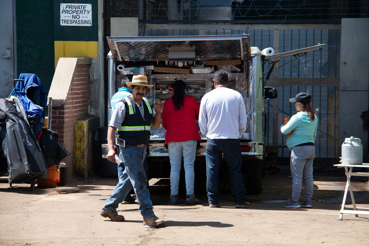 Group of people standing at a food truck in an industrial alley, with a man in a straw hat walking past holding a drink. Group of people standing at a food truck in an industrial alley, with a man in a straw hat walking past holding a drink.