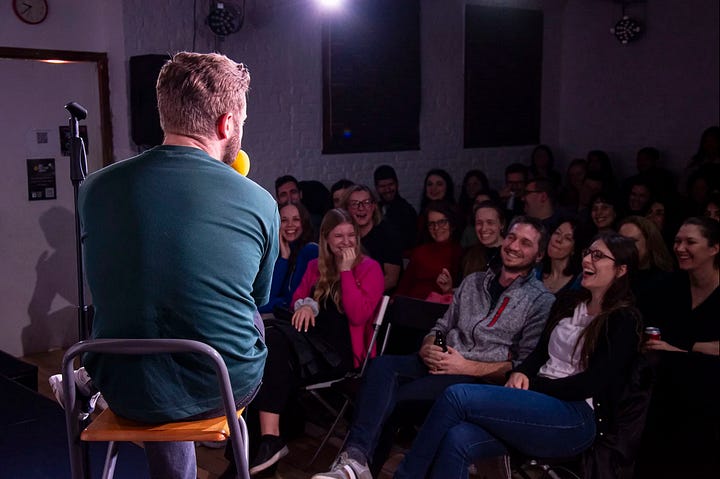 In the left-hand photo, a packed audience watches a performer off-camera in a low-lit bar in the evening. They are applauding. In the right-hand photo, a man sits on a stool on a stage with his back turned to the camera speaking into a mic to a listening audience. The audience are laughing.