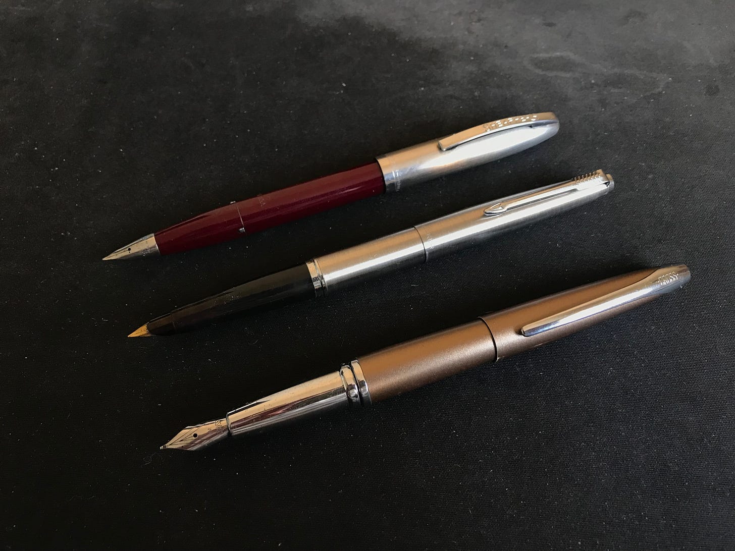 Three fountain pens on a desk. A 1960s Sheaffer, a 1979 Parker and an early 2000s Cross. Three fountain pens on a desk. A 1960s Sheaffer, a 1979 Parker and an early 2000s Cross.