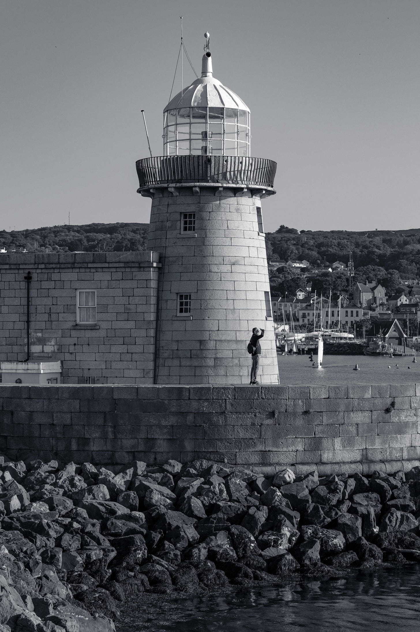 A person stands below the stone lighthouse at Howth Harbour, looking up toward its glass dome in the late afternoon light. A person stands below the stone lighthouse at Howth Harbour, looking up toward its glass dome in the late afternoon light.