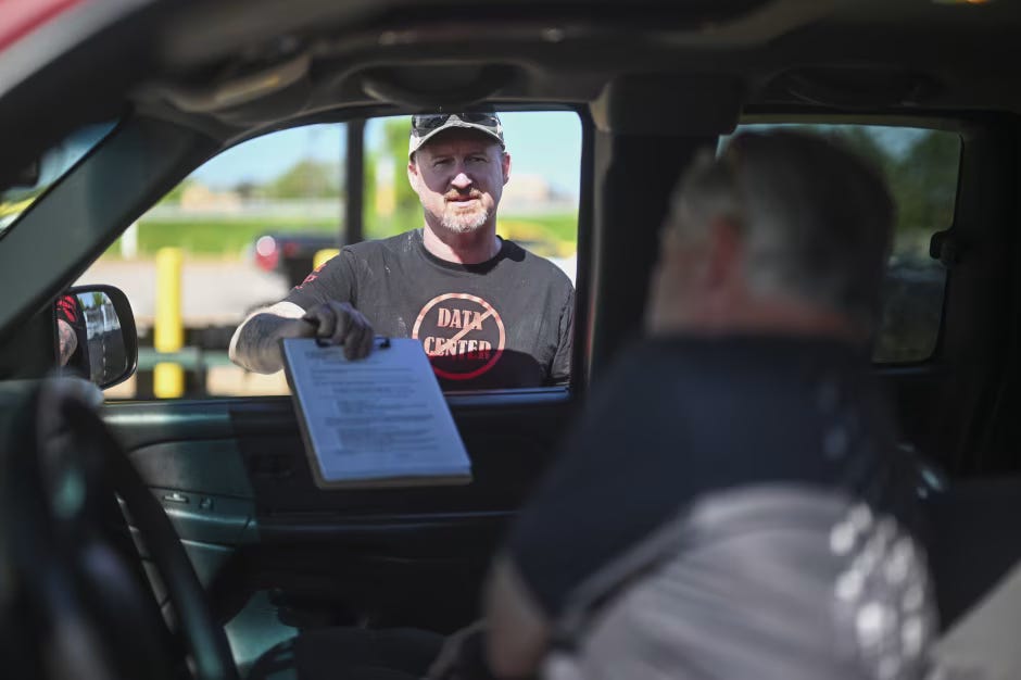 Gabriel Cotton, wearing a "DATA CENTER" t-shirt, holds out a clipboard with a complaint form to a person inside a vehicle. Gabriel Cotton, wearing a "DATA CENTER" t-shirt, holds out a clipboard with a complaint form to a person inside a vehicle.