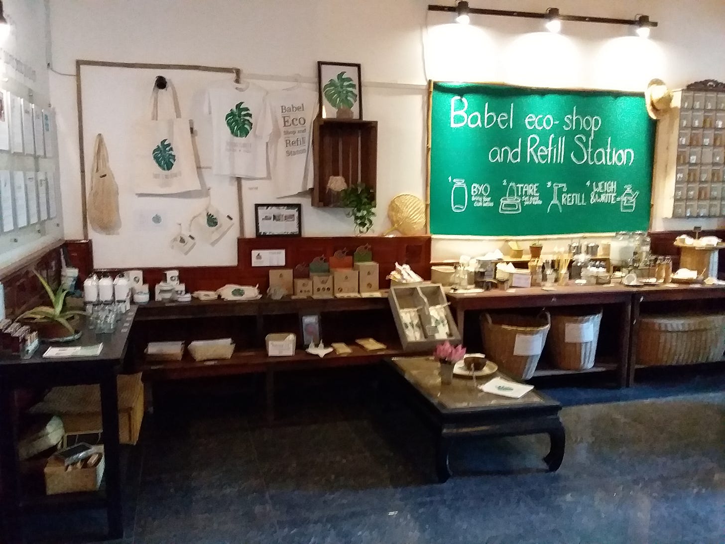 A photograph of a zero-waste store corner titled "Babel eco-shop and Refill Station." A large green chalkboard with white writing illustrates a step-by-step refill process. Below the board, wooden tables and shelves display various eco-friendly products, glass jars, and large woven baskets. To the left, white tote bags and t-shirts featuring a green monstera leaf logo hang on the wall.