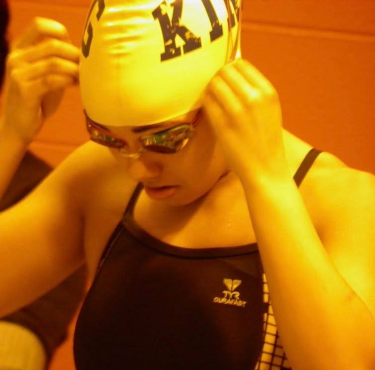Orange-toned photo of teen in swimsuit and swimcap adjusting goggles