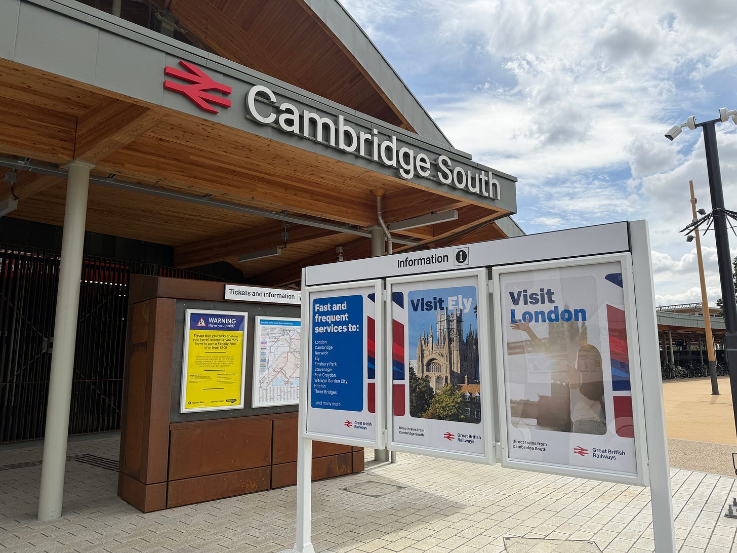 Photo of western entrance to Cambridge South station showing station name signage, double arrows, and GBR branded posters.