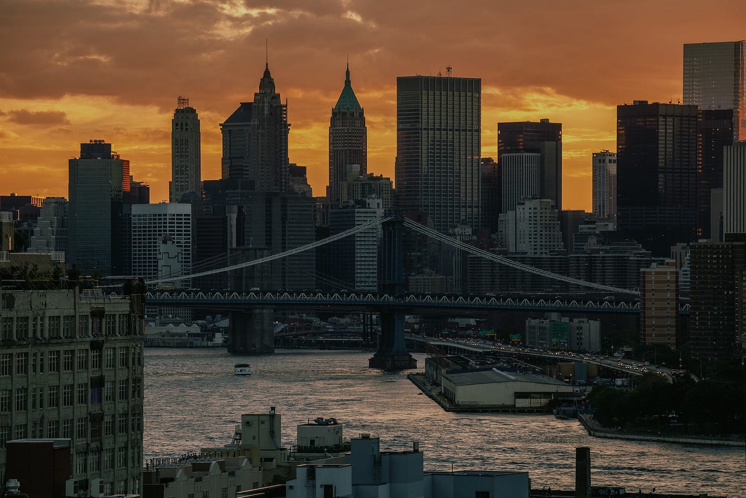 An elevated view of the Lower Manhattan skyline and the Manhattan Bridge at sunset. The sky is a dramatic orange, and the buildings are silhouetted against it, with the East River in the foreground.