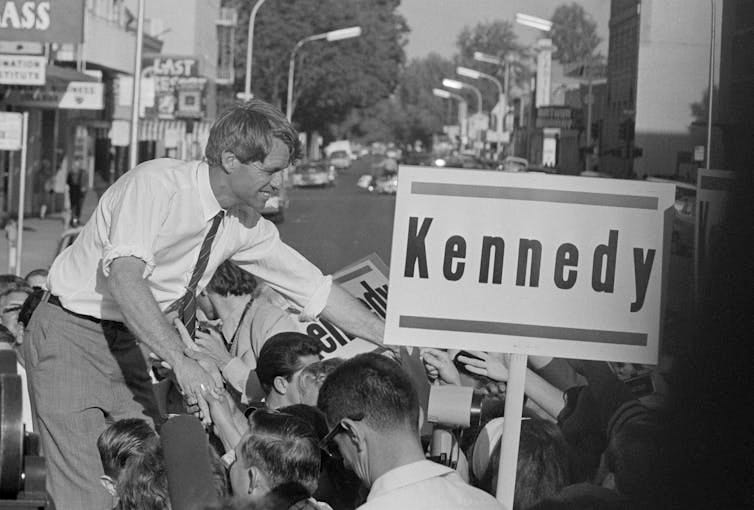 A man in shirtsleeves standing on a car, reaching out to shake people's hands. A man in shirtsleeves standing on a car, reaching out to shake people's hands.
