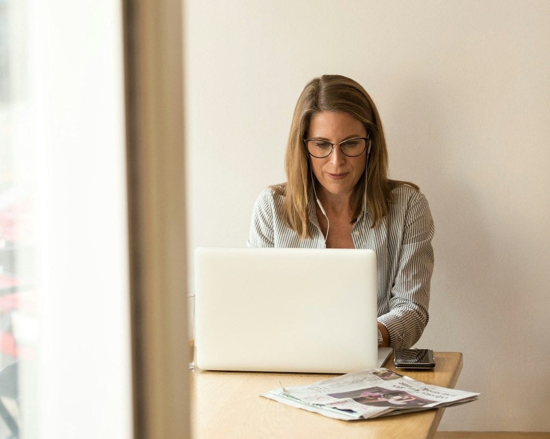 woman wearing grey striped dress shirt sitting down near brown wooden table in front of white laptop computer