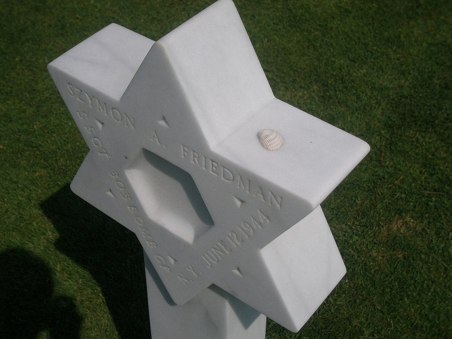 A white marble Star of David headstone bearing Szymon A. Friedman’s name at the Normandy American Cemetery, with a small seashell resting on it. A white marble Star of David headstone bearing Szymon A. Friedman’s name at the Normandy American Cemetery, with a small seashell resting on it.