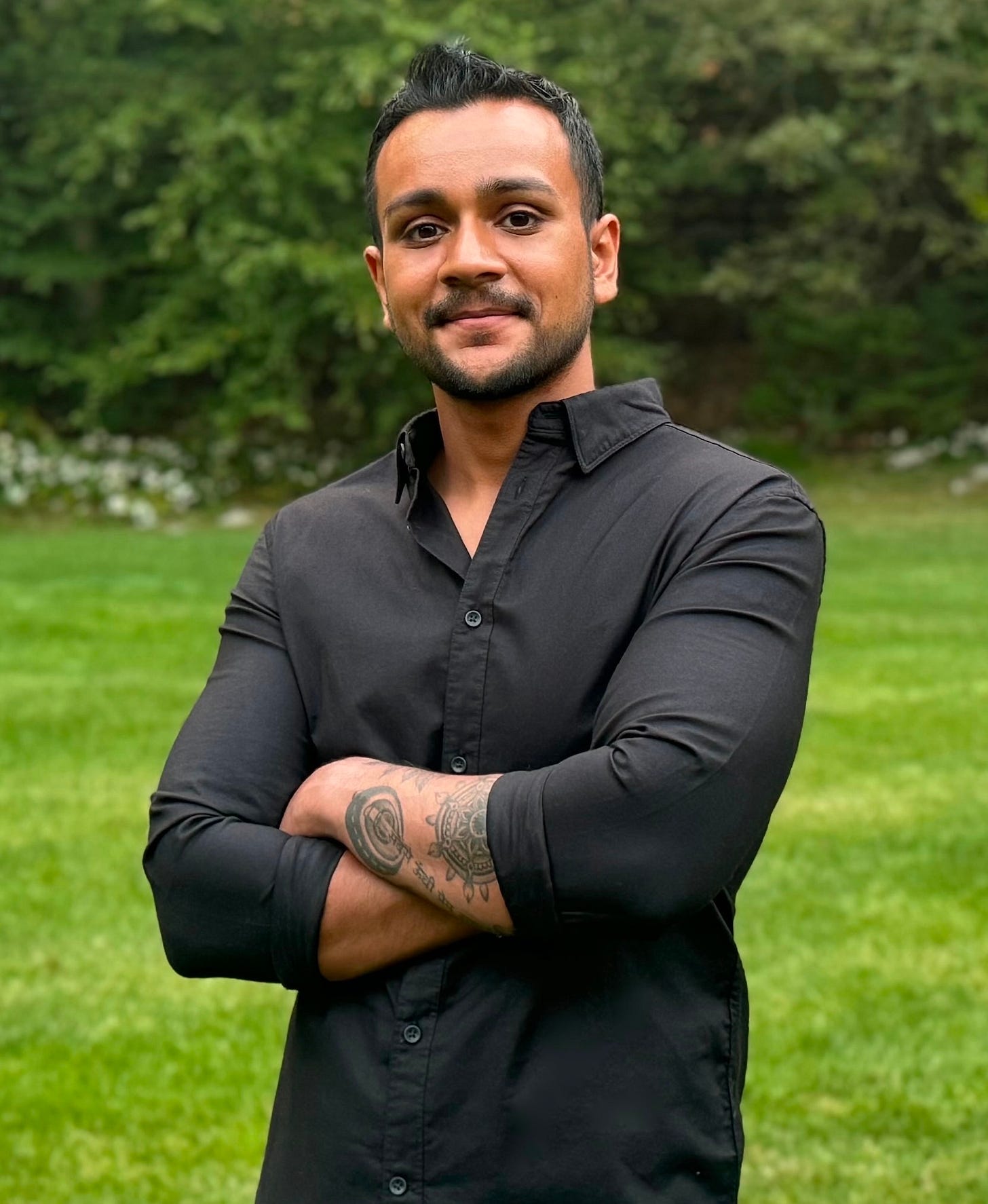 An adult man is centered in the frame, facing the camera with his arms crossed and a slight smile. He wears a black button-up shirt, with tattoos visible on one forearm. The background is softly blurred, showing a green lawn and dense trees that create strong contrast with the dark clothing.