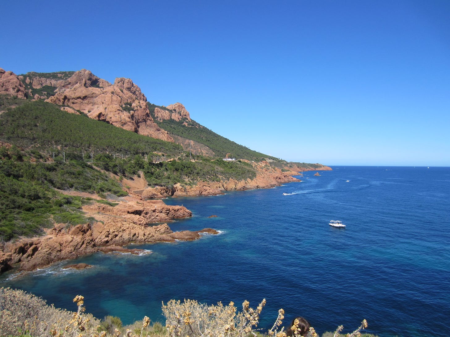 The bright blue water of the Mediterranean with a perfectly clear sky above it. To the left is a rocky red hillside partially covered in green vegation