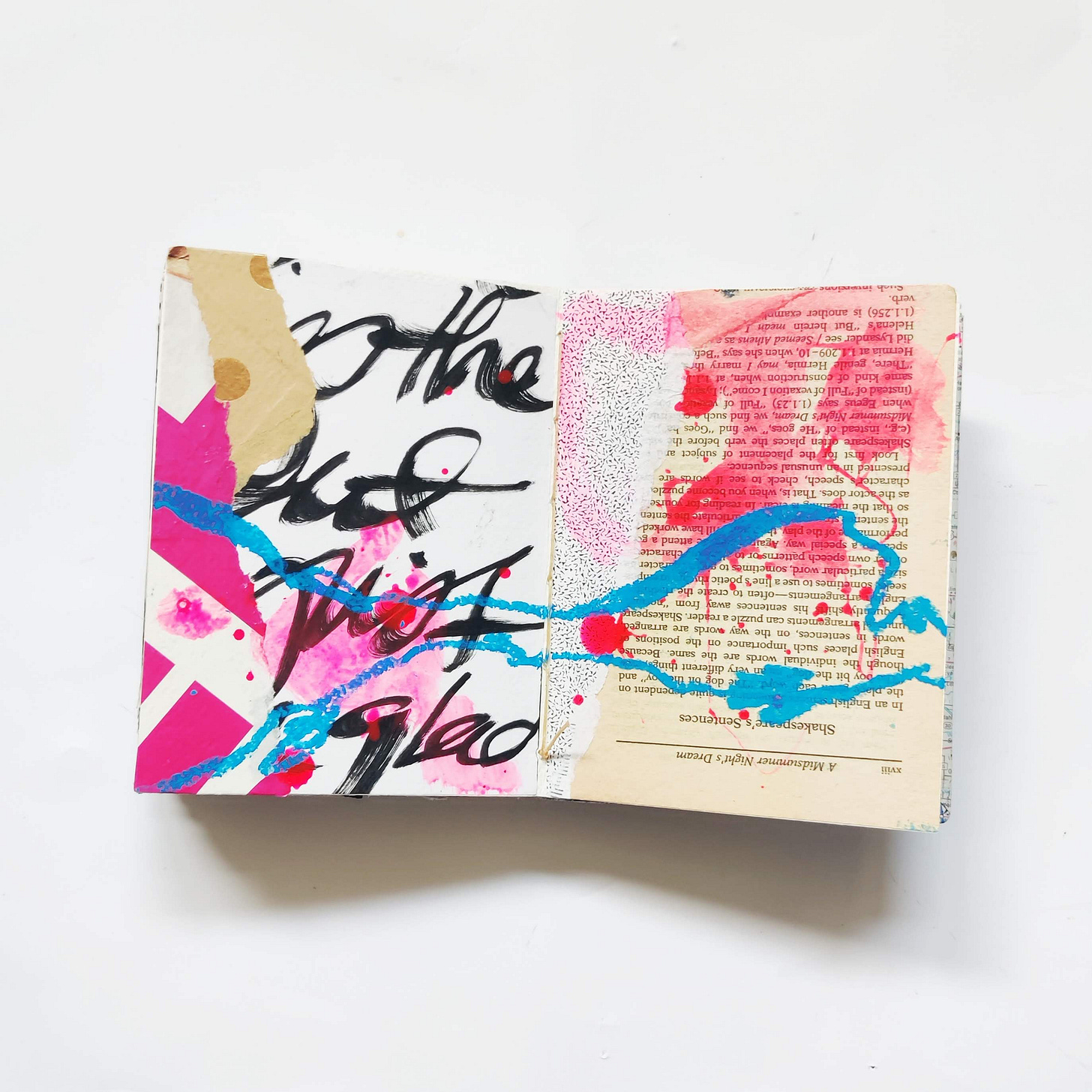 Open mixed media art journal. A layer of collage — an aged book page, large black handwriting, and pink and brown papers — are topped with horizontal scribbles in blue oil pastel and splatters of bright pink ink splatters. Open mixed media art journal. A layer of collage — an aged book page, large black handwriting, and pink and brown papers — are topped with horizontal scribbles in blue oil pastel and splatters of bright pink ink splatters.