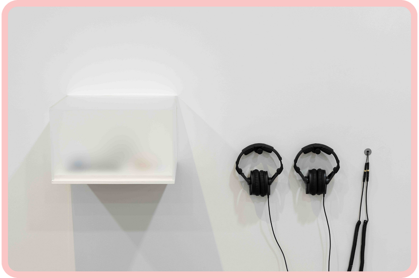 A photo of an opaque glass vitrine mounted on a white wall. To its right, a set of black headphones are also attached to the wall. A photo of an opaque glass vitrine mounted on a white wall. To its right, a set of black headphones are also attached to the wall.