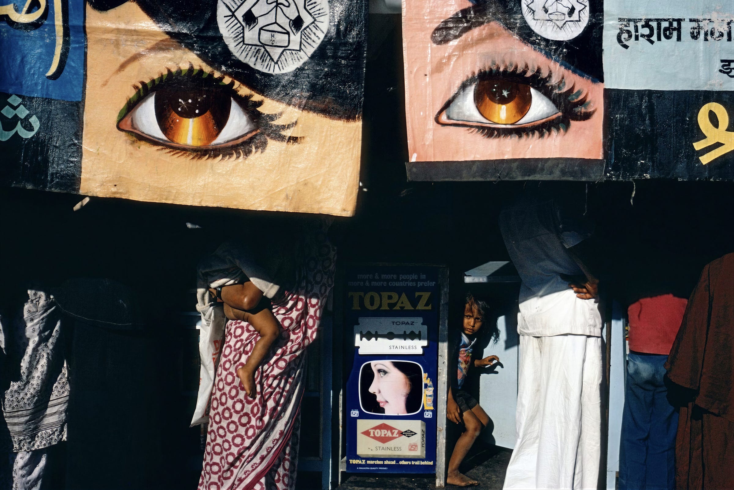 Alex Webb - Colorful painted banners with large eyes hang above a stall. People in patterned clothing walk by and through the stall, while a child peeks out from behind a display advertising TOPAZ with an image of a womans face.