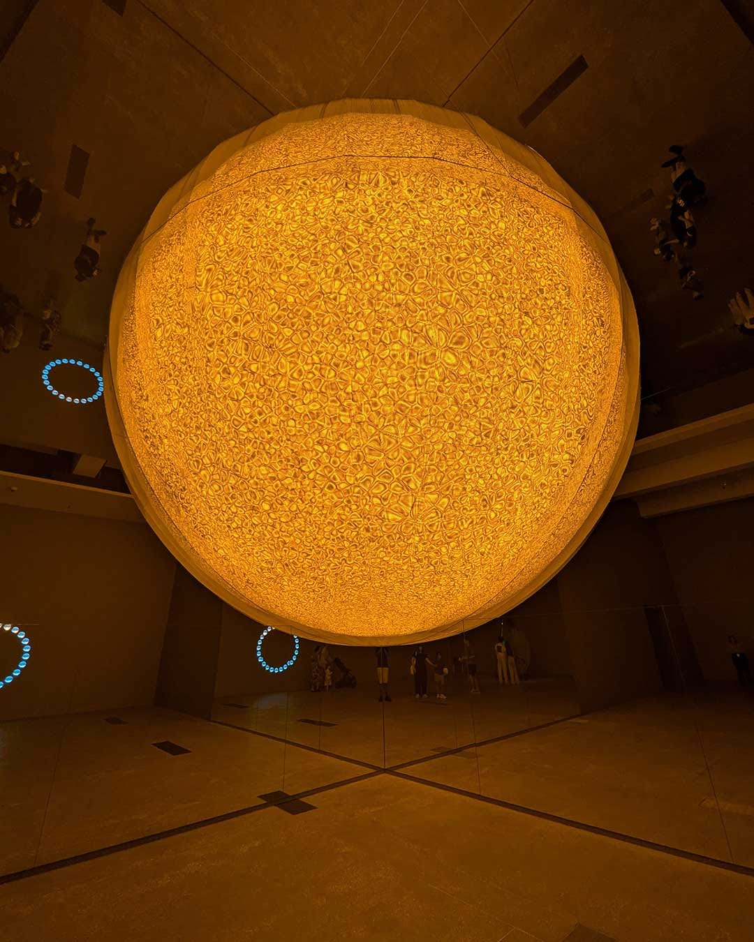 View looking directly upward at large circular installation with intricate geometric pattern illuminated in warm golden-yellow light against dark gallery ceiling, with silhouettes of visitors visible at edges