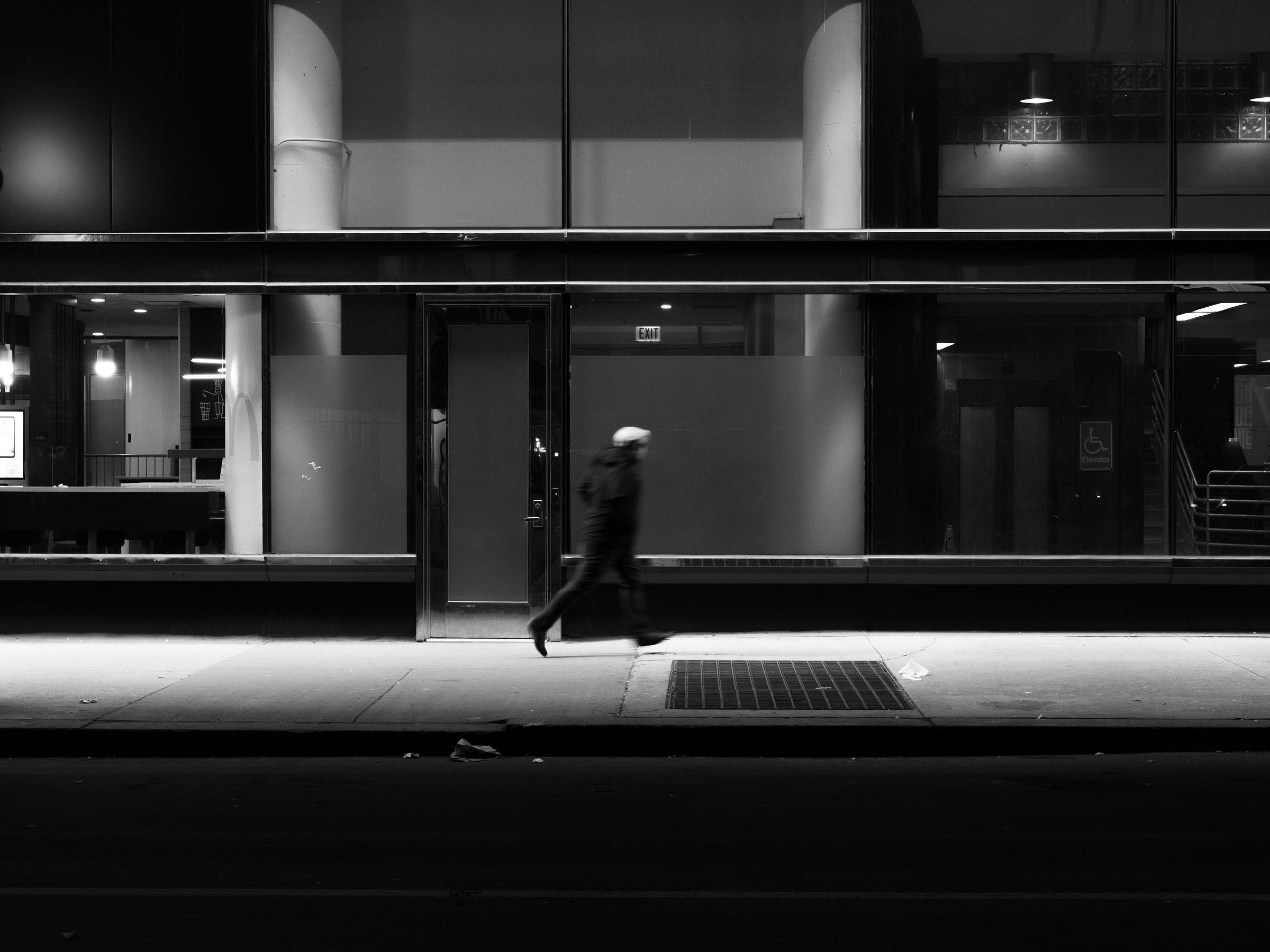 Black and white photograph of a solitary figure walking past a modern glass building at night. The person, wearing a hat and carrying a bag, moves briskly through the illuminated sidewalk, their blurred motion contrasting with the still, empty interior of the building behind them. Black and white photograph of a solitary figure walking past a modern glass building at night. The person, wearing a hat and carrying a bag, moves briskly through the illuminated sidewalk, their blurred motion contrasting with the still, empty interior of the building behind them.