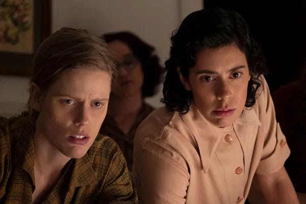 Two serious-looking women are leaning forward as they look slightly off screen. The pale woman on the left has dark blonde hair pulled back in a low braid and is wearing an olive green plaid shirt. The woman on the right has shoulder-length dark curly brown hair and is wearing a beige button-up shirt with pointy collars.