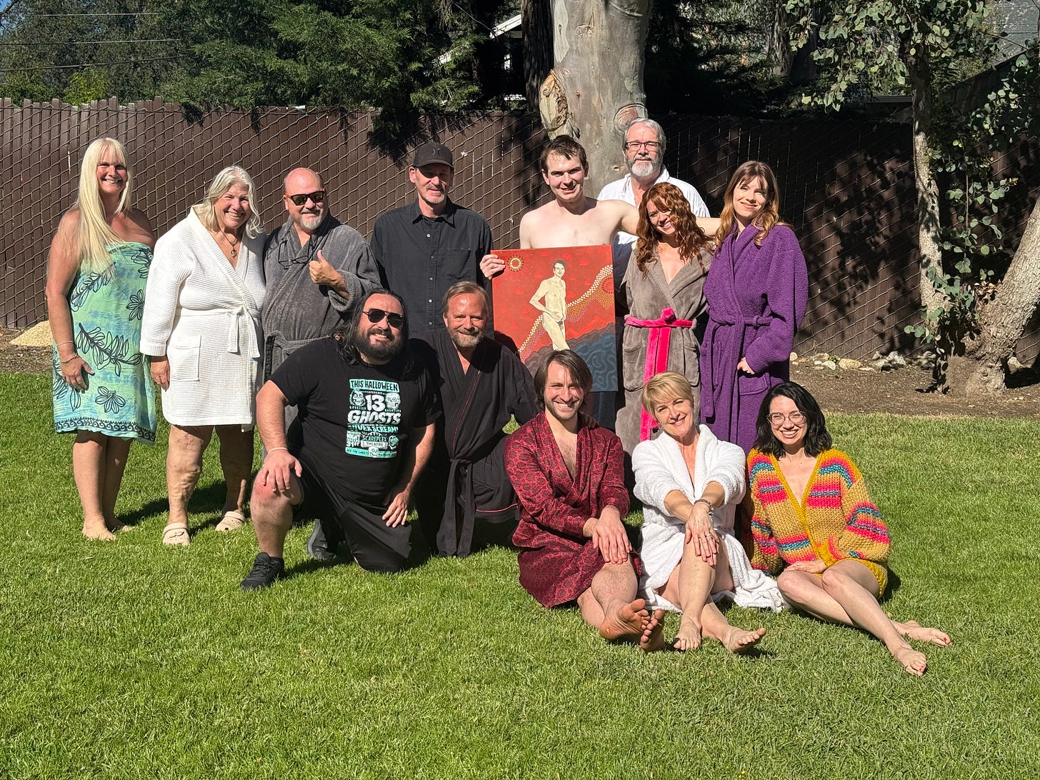 A group of cast and crew pose together on a sunlit lawn for a photo, many wearing robes despite the film’s clothing-optional setting. Some stand while others sit in front. At left, Linda Weber, Cynthia Holbach, and Rolf Holbach stand together, with trees and a fence in the background.