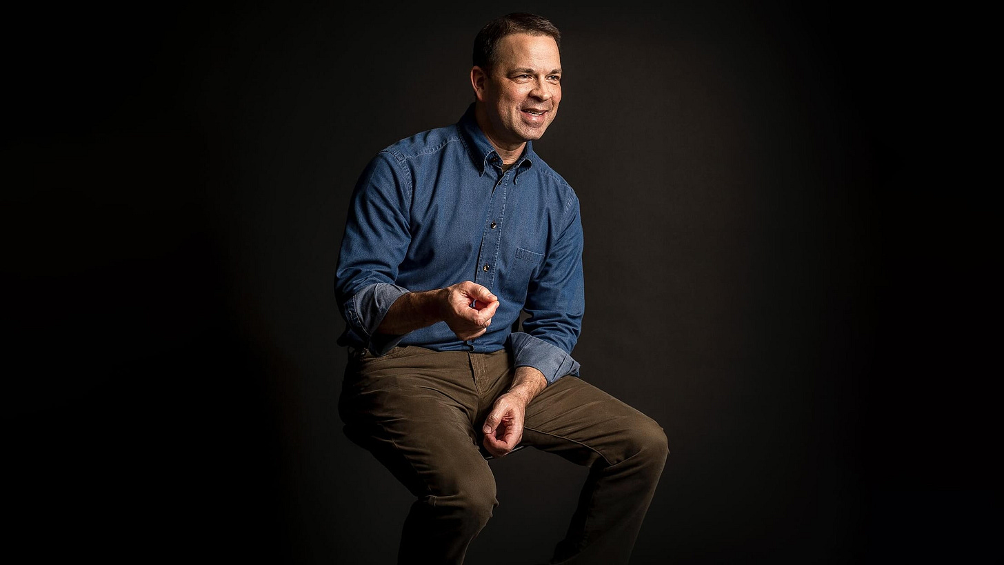 A photo of Dieste CEO Greg Knipp smiling as he's sitting on a stool and looking off to the right, while the background is a light brownish gray color that fades to black.