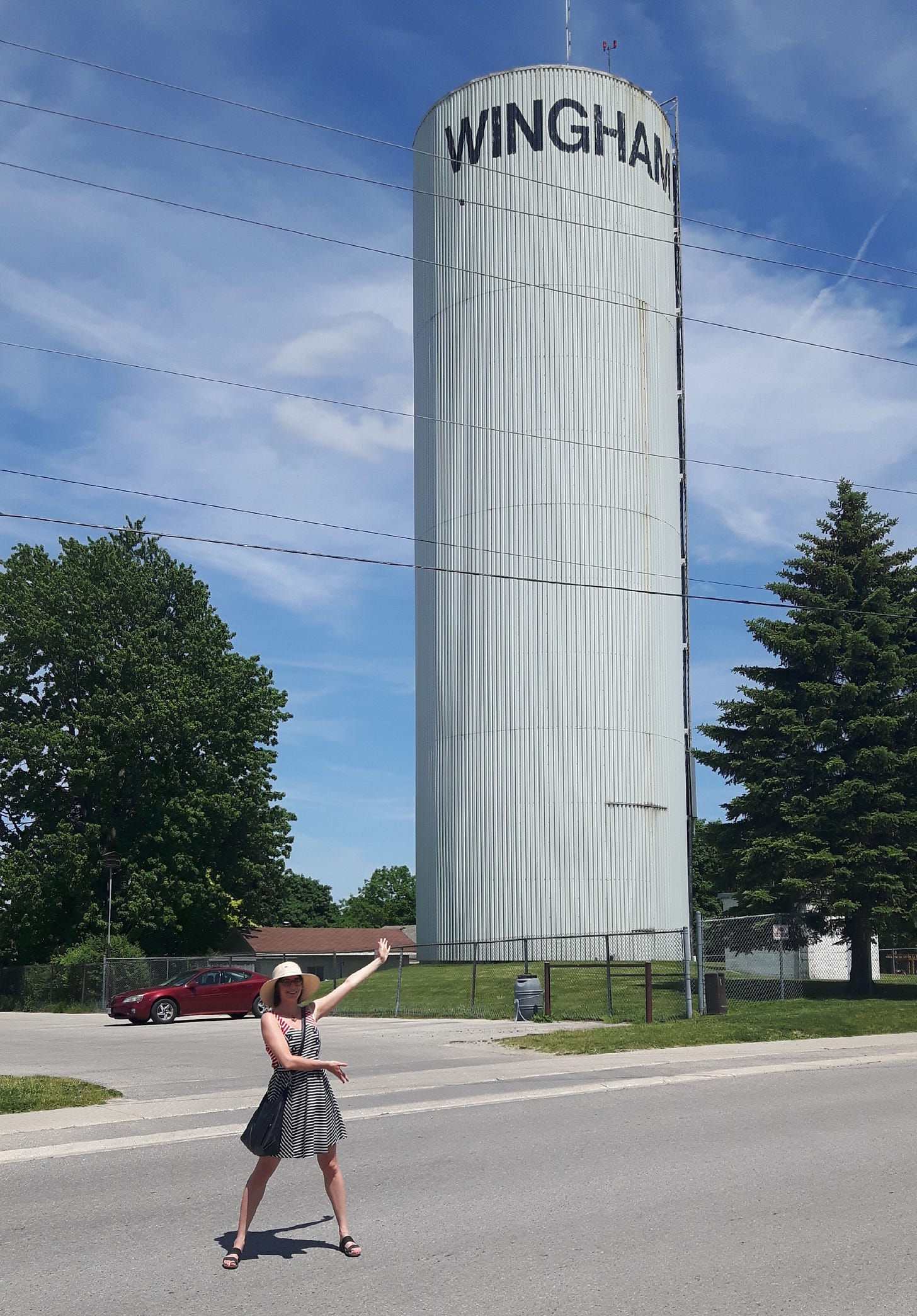 Kathy Friedman wearing a white sunhat and black and white dress standing with her arms up in front of the Wingham water tower.