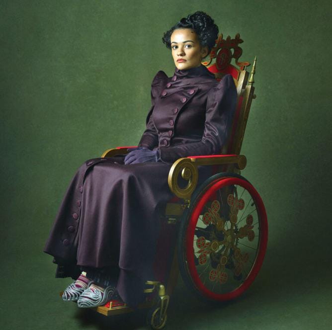 In a photo set against a green background, Nessa is wearing a fancy, deep purple overcoat and silver slippers. Her wheelchair is ornate with red wheels and curved golden flourishes.