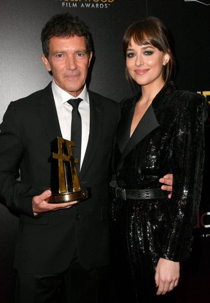 Two people dressed in formal black clothing pose for a photo. One holds a film award trophy Two people dressed in formal black clothing pose for a photo. One holds a film award trophy
