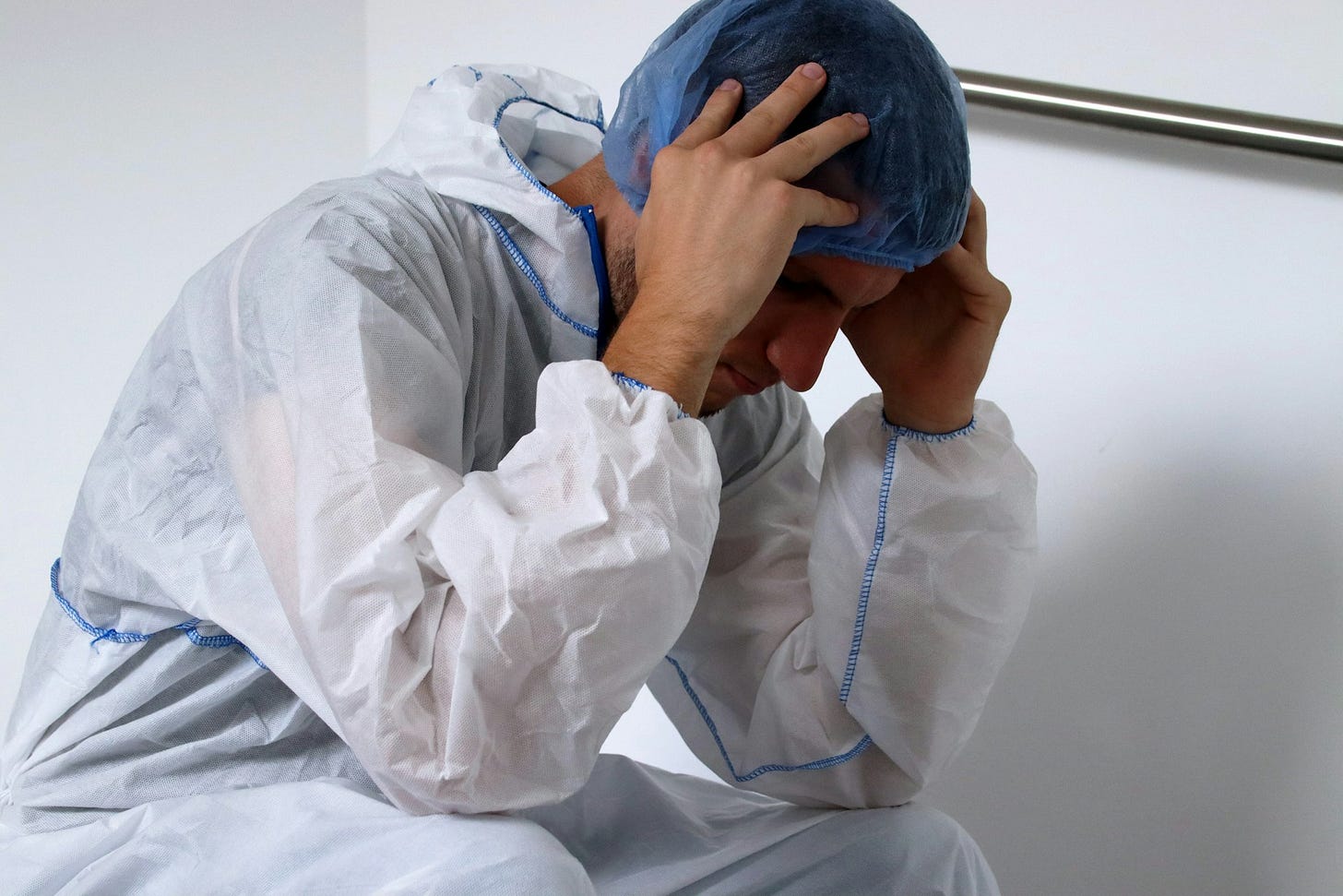 A fatigued health care worker in protective clothing sits with head bowed and hands on temples, showing exhaustion during a hospital shift. Photo by Maskmedicare Shop on Unsplash