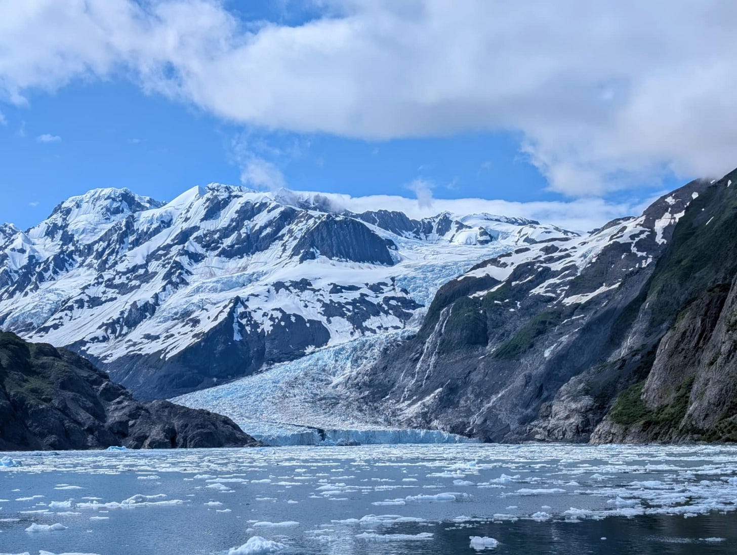 A stunning glacier flows between snow-covered mountains into an icy, blue-gray bay scattered with floating ice chunks, under a bright sky with scattered clouds.  