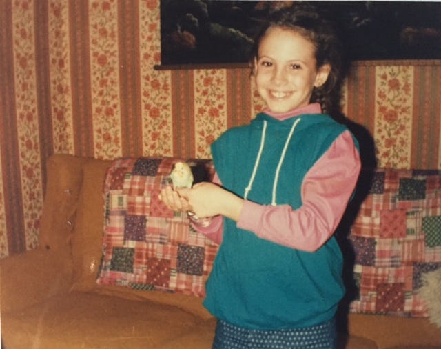 Me as a kid, hair in two french braids, wearing a multi-colored sweatshirt, and holding a paraket in a 70s styled living room..
