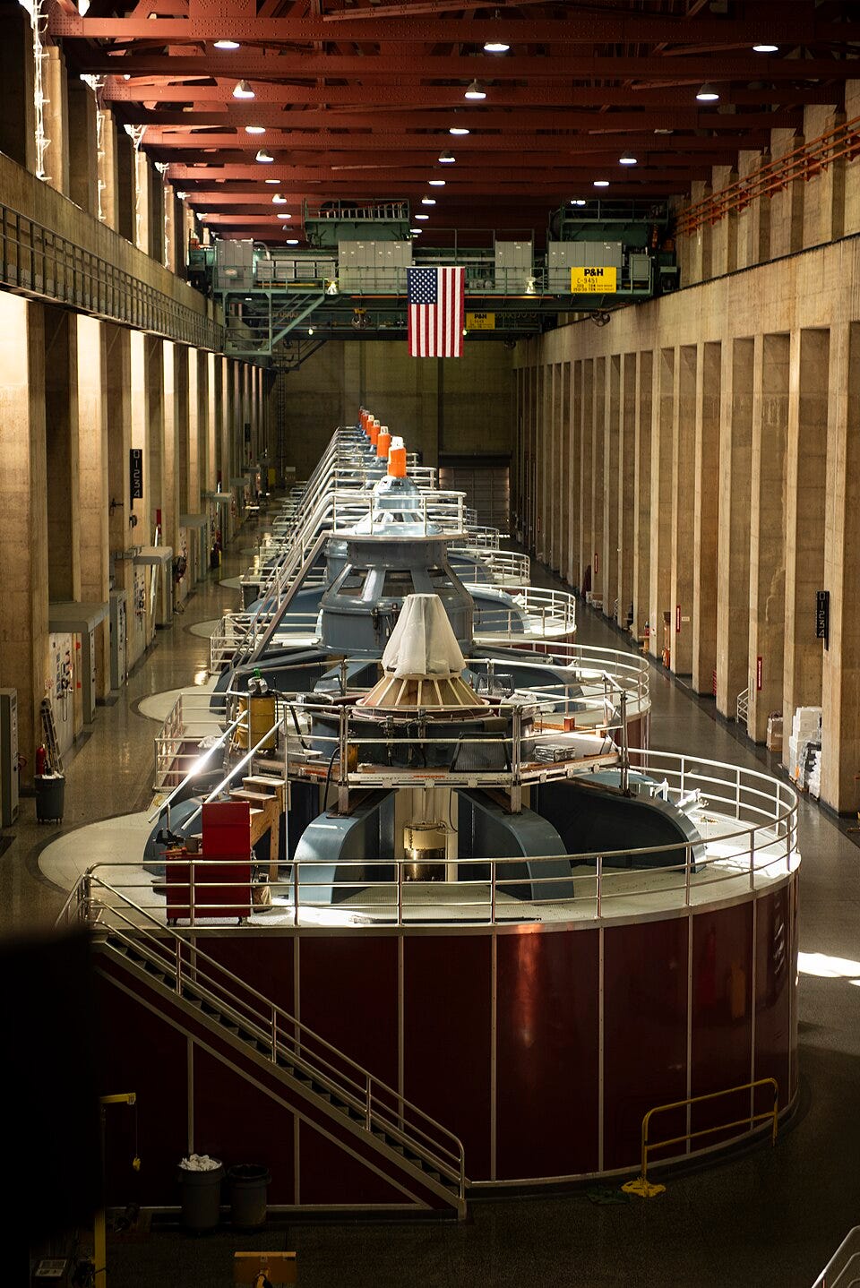A picture looking down on eight generators in a line receding away from the viewer. Each generator is a cylinder with dark red walls, with a railing and walkable space on the white top of each generator. On top of each generator is a blue structure that comes to a point. On the very top of each generator is are white lights with black letters "N1" (closest) through "N8" (furthest). All are lit except for "N4". There are access catwalks on top of each generator. The generators are in a large stone hallway with metal beams in the ceiling. There are two gantry cranes at the far end of the room. A picture looking down on eight generators in a line receding away from the viewer. Each generator is a cylinder with dark red walls, with a railing and walkable space on the white top of each generator. On top of each generator is a blue structure that comes to a point. On the very top of each generator is are white lights with black letters "N1" (closest) through "N8" (furthest). All are lit except for "N4". There are access catwalks on top of each generator. The generators are in a large stone hallway with metal beams in the ceiling. There are two gantry cranes at the far end of the room.