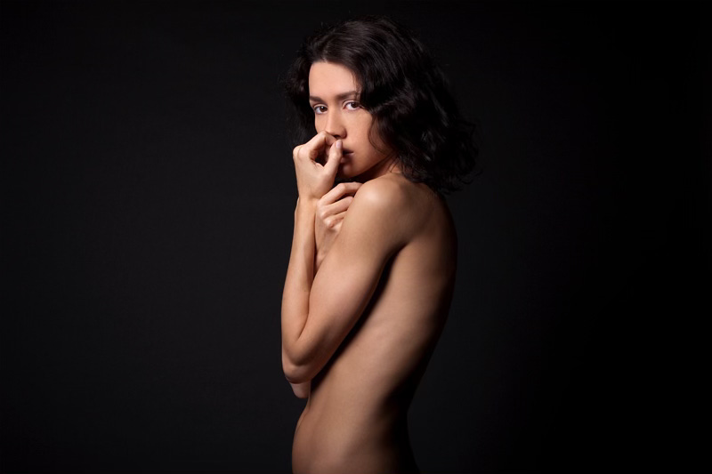Ritratto di una giovane donna su fondo nero. La sua pelle nuda risalta ed emerge dal fondo scuro. L'espressione è dolce, quasi intimorita. Con una mano copre delicatamente la bocca, come in un gesto di protezione