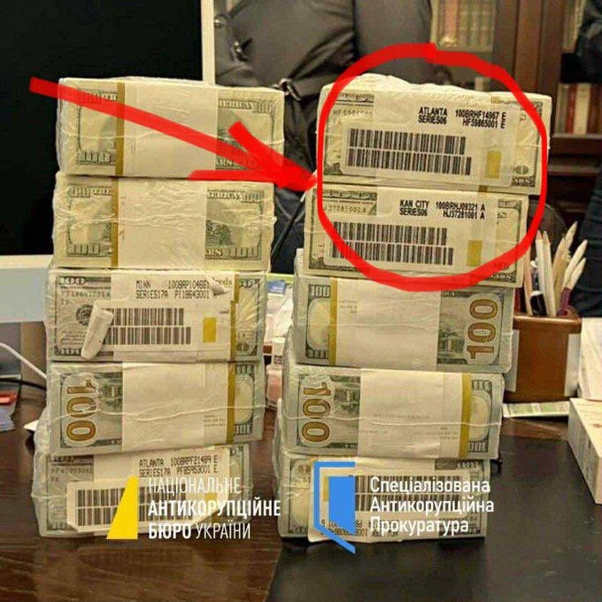 Stacks of bundled US dollar bills in clear plastic wrapping on a wooden desk with office supplies including pens and papers nearby. Red arrows highlight the packaging labels marked Atlanta and Kansas City from the Federal Reserve. Ukrainian text overlays indicate anti-corruption committee involvement.