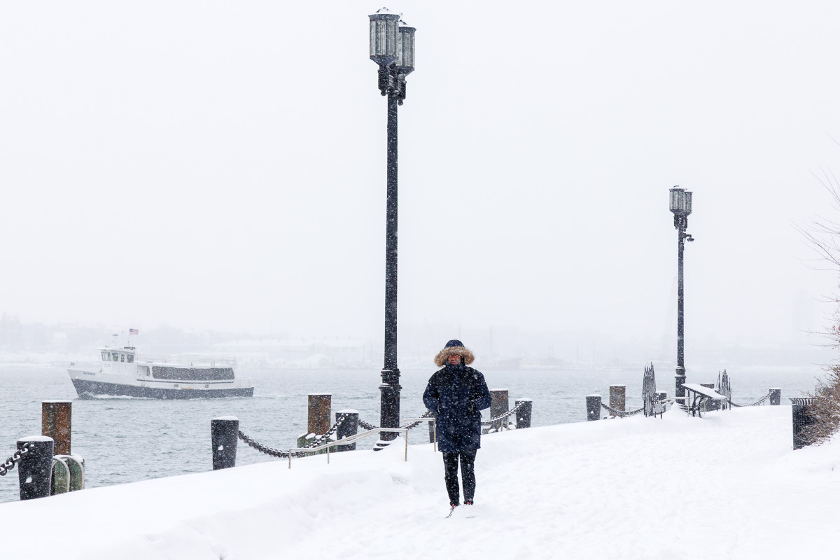 Person walking along the snowy HarborWalk with a ferry passing in the background during a snow squall.