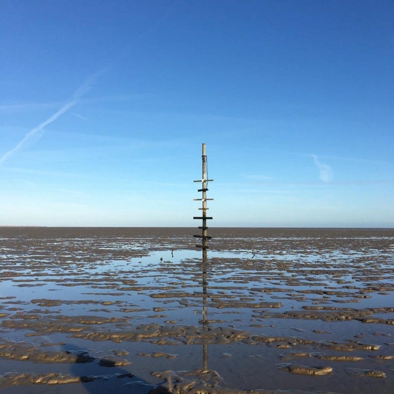 A “maypole” stands as a marker point on The Broomway off Foulness Island, Essex, a perilous path that disappears when the tide comes in. The sky is clear with just a few contrails. The blue sky reflects off the dabbled shallow water.
