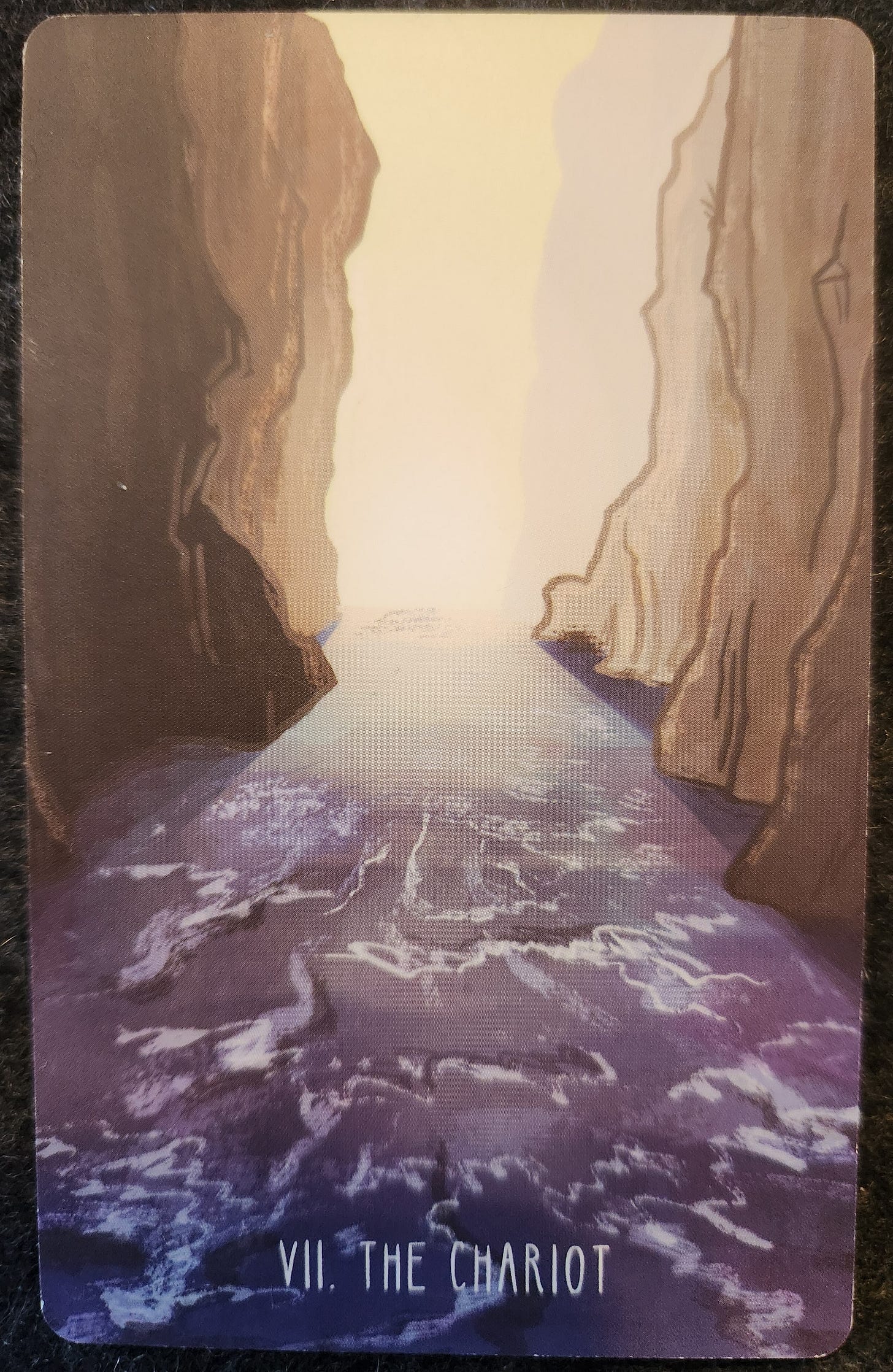 A tarot card labelled VII The Chariot. It shows a rapidly moving river between two tall cliff faces. The river moves into the distance and appears to emerge from the cliffs. The distant sky is bright with sunlight. 