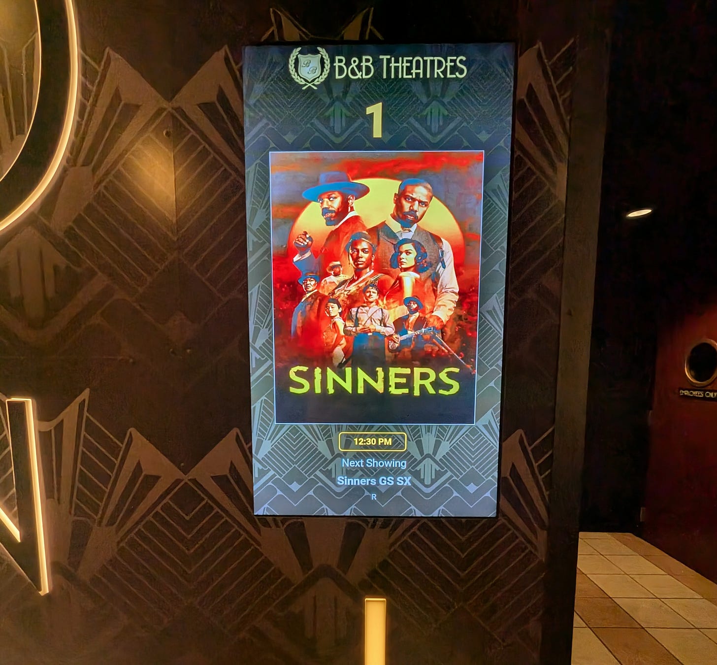 A picture of the digital marquee next to a movie theater. The digital poster reads SINNERS 12:30pm Next Showing Sinners GS SX R. The display is on a brown and gold wall with art deco style wallpaper. A brown and white tiled floor to the right of the marquee leads into the theater.