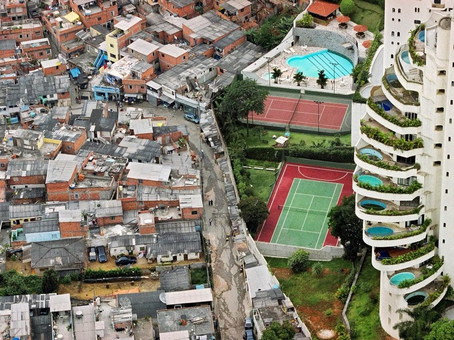 Aerial view shows a densely packed slum neighborhood with numerous small brick houses clustered together many with red-tiled roofs and some blue accents narrow dirt roads winding between them and scattered vegetation on one side while on the other side a tall white high-rise apartment building with curved balconies and green landscaping stands adjacent to a rectangular blue swimming pool a red tennis court surrounded by grass and palm trees and additional green spaces. Aerial view shows a densely packed slum neighborhood with numerous small brick houses clustered together many with red-tiled roofs and some blue accents narrow dirt roads winding between them and scattered vegetation on one side while on the other side a tall white high-rise apartment building with curved balconies and green landscaping stands adjacent to a rectangular blue swimming pool a red tennis court surrounded by grass and palm trees and additional green spaces.