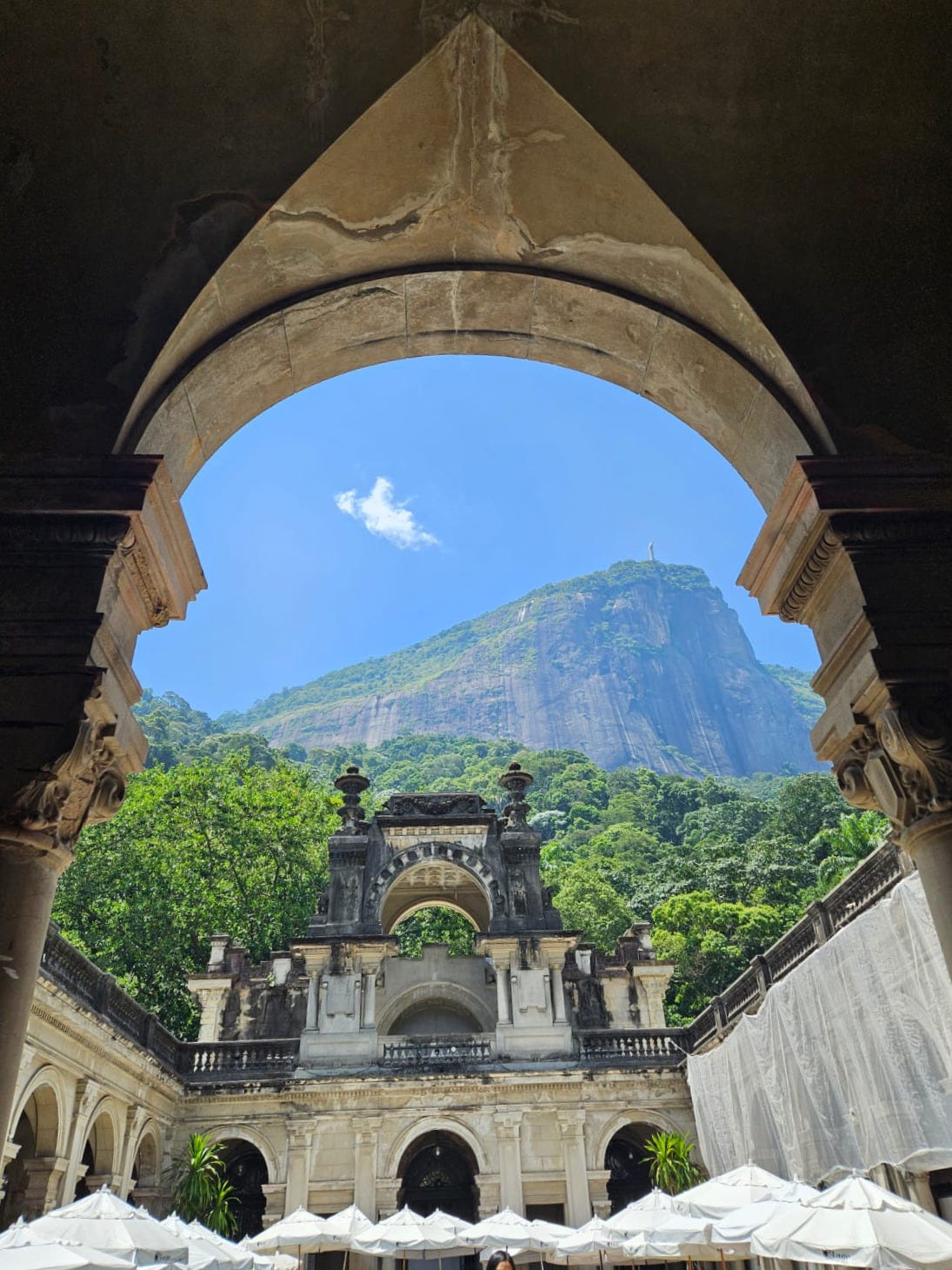 foto tirada de dentro de uma palacete, olhando para cima e para fora. a estrutura do prédio é branca e envelhecida por pontos escuros, imponente, ornamentada. Vê-se na parte de baixo da foto deiversos guarda-sóis, o topo deles. A cima, vê-se um fundo verde e bonito de árvores e o morro com a estátua do Cristo Redentor, com o céu azul ao fundo. Apenas uma nuvem solitária paira ali foto tirada de dentro de uma palacete, olhando para cima e para fora. a estrutura do prédio é branca e envelhecida por pontos escuros, imponente, ornamentada. Vê-se na parte de baixo da foto deiversos guarda-sóis, o topo deles. A cima, vê-se um fundo verde e bonito de árvores e o morro com a estátua do Cristo Redentor, com o céu azul ao fundo. Apenas uma nuvem solitária paira ali